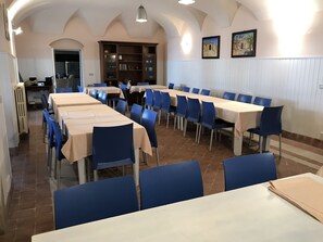 Un comedor con mesas largas y sillas azules, una cocina al fondo, y una pared blanca con dos cuadros.