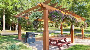 Una pérgola de madera con cestas de flores colgantes, mesas de picnic y una parrilla en un parque.