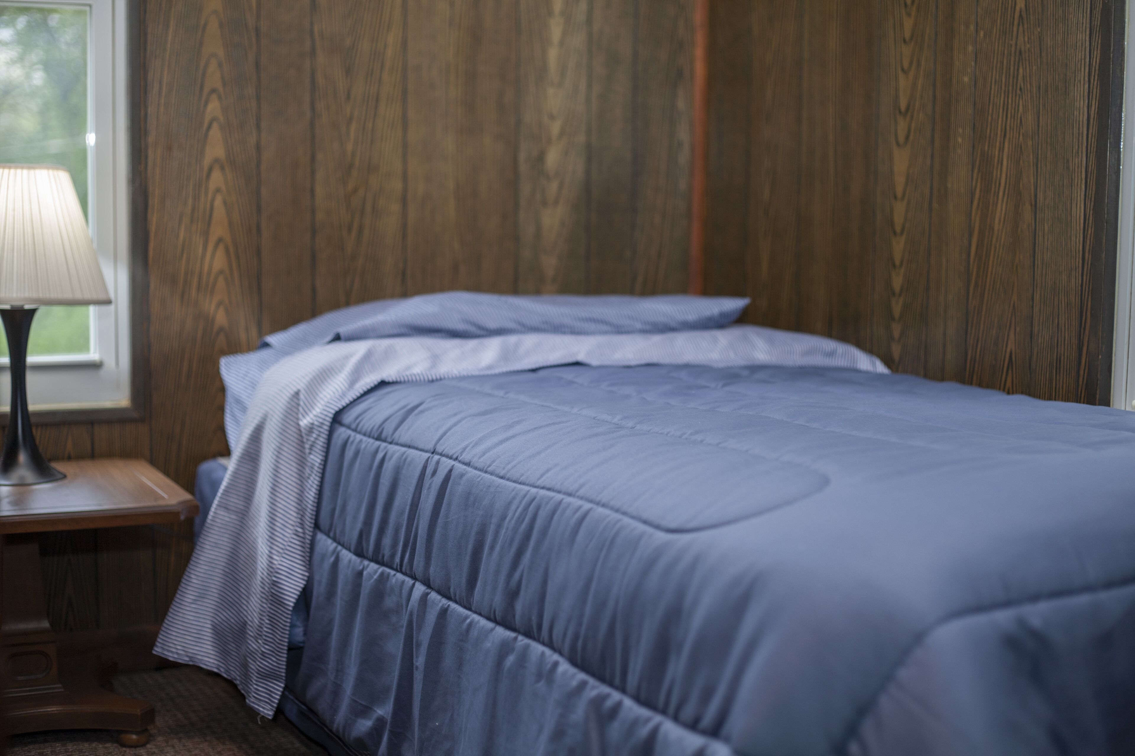 Una cama bien hecha con edredón azul y almohadas, una mesita de noche de madera con una lámpara, y una ventana con vistas a zonas verdes.