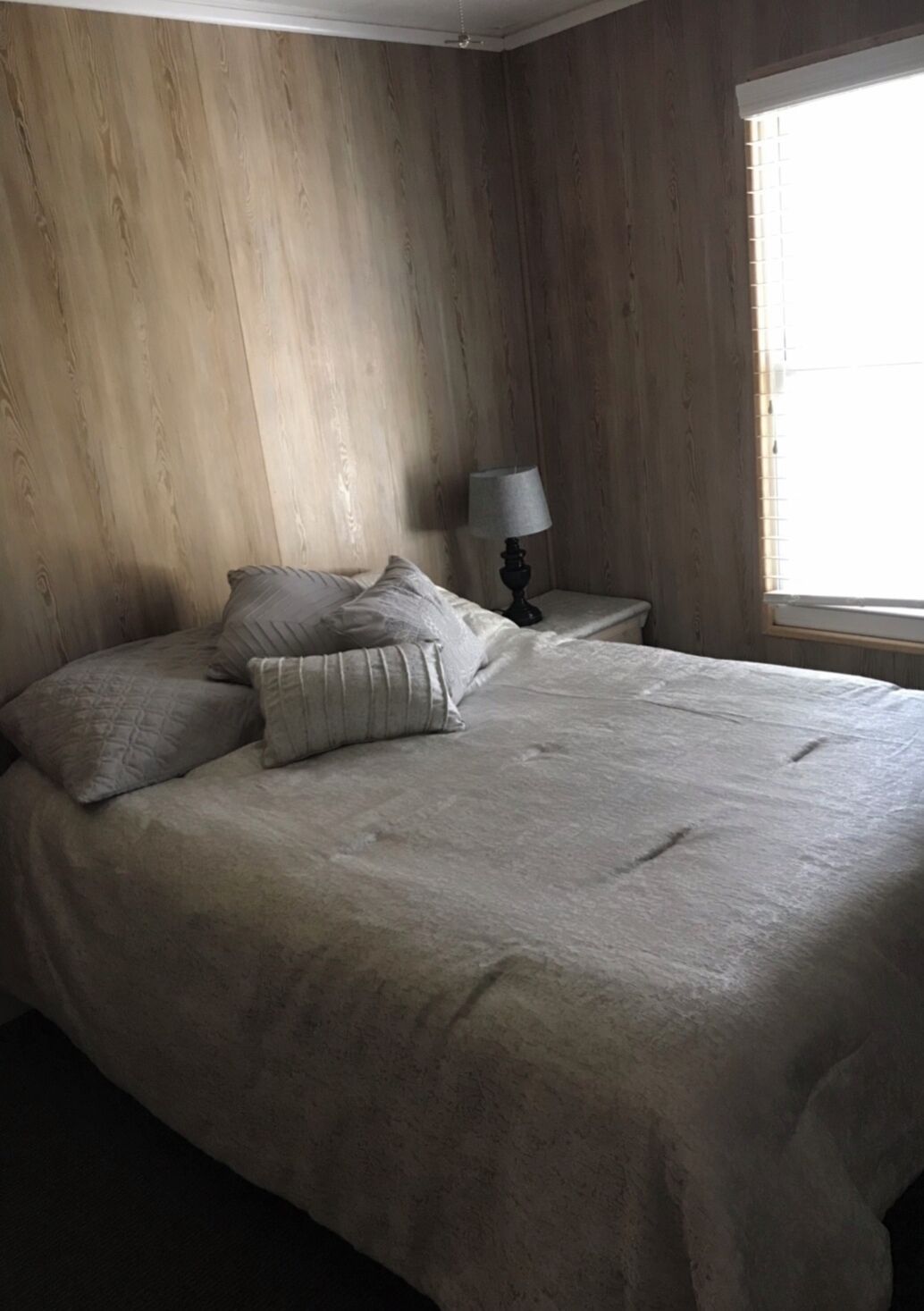 A bedroom with a bed, pillows, a nightstand with a lamp, wooden walls, and a window.