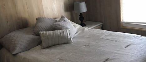 A bedroom with a bed, pillows, a nightstand with a lamp, wooden walls, and a window.