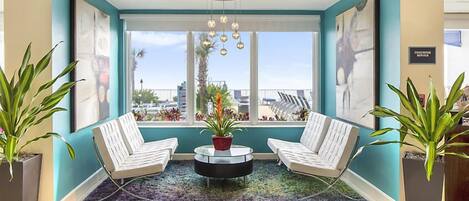 Un vestíbulo moderno con dos sillones blancos, una mesa redonda para café y un ventanal con vistas a palmeras y edificios.