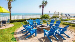 Un patio frente a la playa con sillas Adirondack azules, un pozo de fuego y un paraguas amarillo.