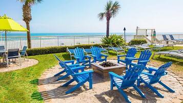 Une terrasse au bord de la plage, avec des fauteuils Adirondack bleus, un foyer et un parasol jaune.