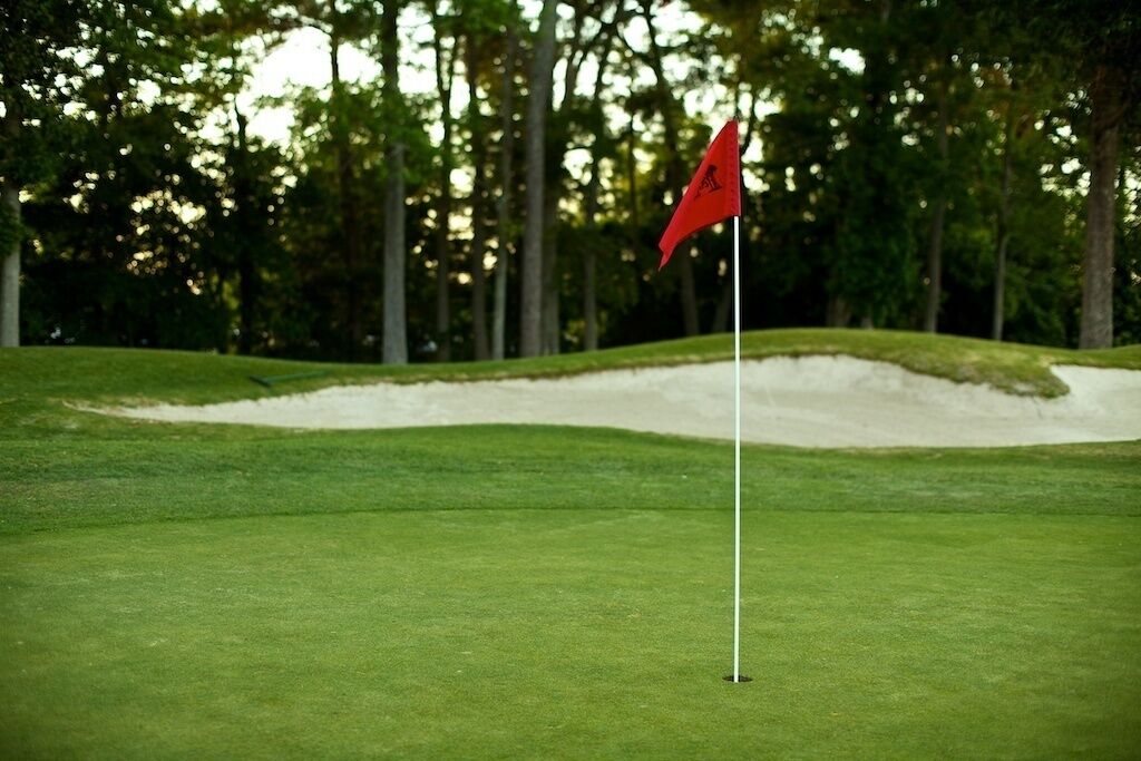 A golf course with a red flag on a white pole, surrounded by green grass and trees.