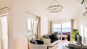 A spacious living room with a large window offering a view of the sea, a white sofa set, a white armchair, and a modern pendant light.