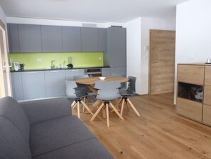 A compact living space with a grey sofa, a wooden dining table with four chairs, a kitchenette with grey cabinets and a green backsplash, and a wooden wardrobe.
