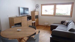 Ein stilvolles Wohnzimmer mit einem Holzesstisch, blauen Stühlen, einem grauen Sofa, einem Fernseher auf einer Holzkommode und einem großen Fenster mit Aussicht.