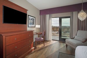 A hotel room with a flat-screen TV, a wooden dresser, a chair, a sofa, a balcony with a view, and a chandelier.