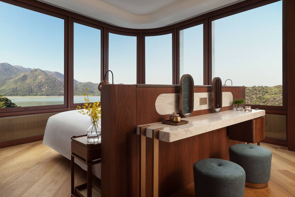 Een moderne hotelkamer met een groot bed, een houten bureau, twee voetenbankjes en uitzicht op bergen door grote ramen.