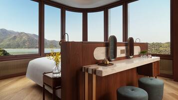 Una habitación de hotel moderna con una cama grande, un escritorio de madera, dos otomanas y vistas a montañas a través de amplios ventanales.