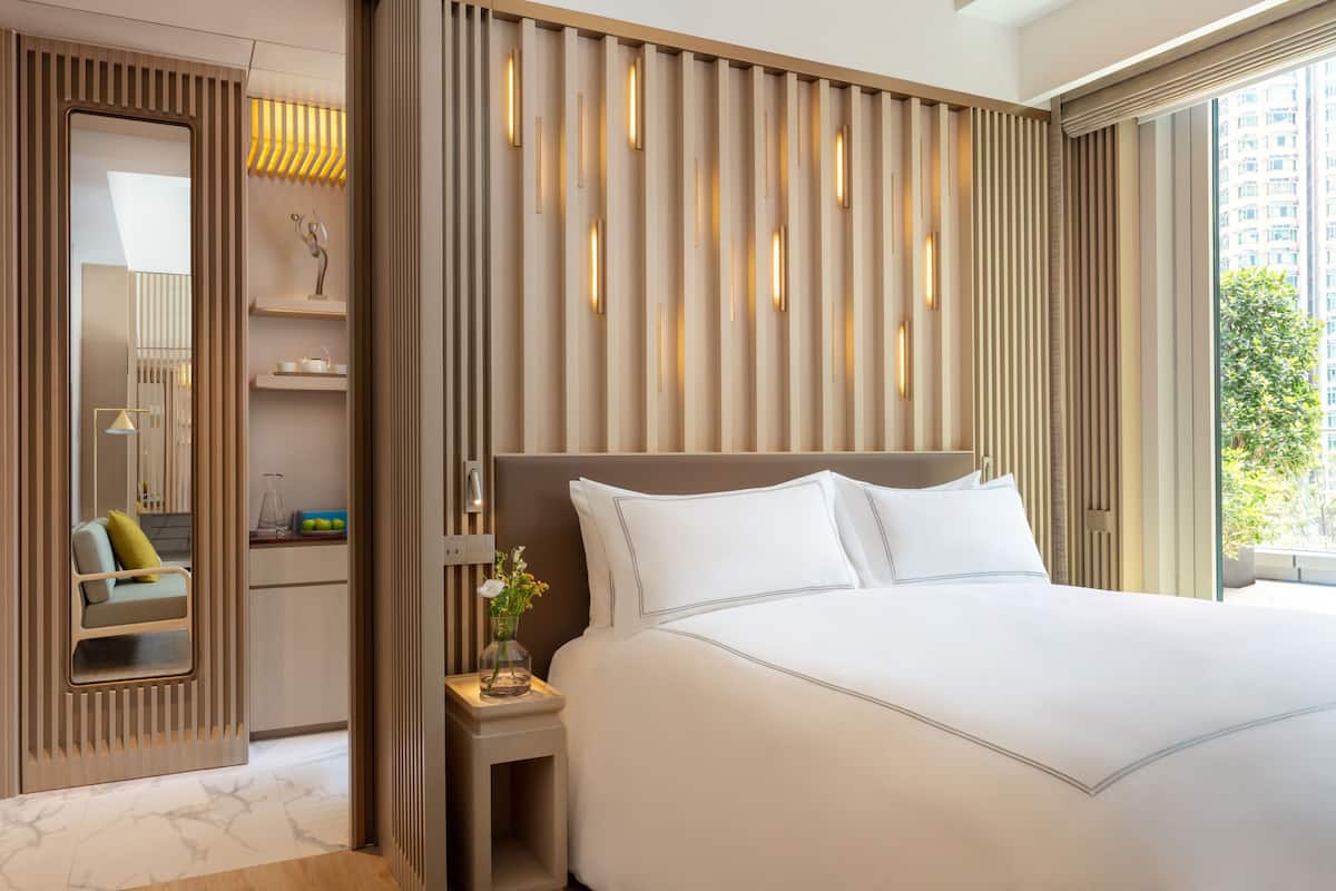 Een moderne hotelkamer met een groot bed, een nachtkastje met een bloevas, een inbouwkast en een groot raam met uitzicht op de stad.