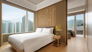 Una habitación de hotel moderna con una cama grande, un panel de madera decorativo, una pequeña mesa con un florero y vistas a edificios altos.