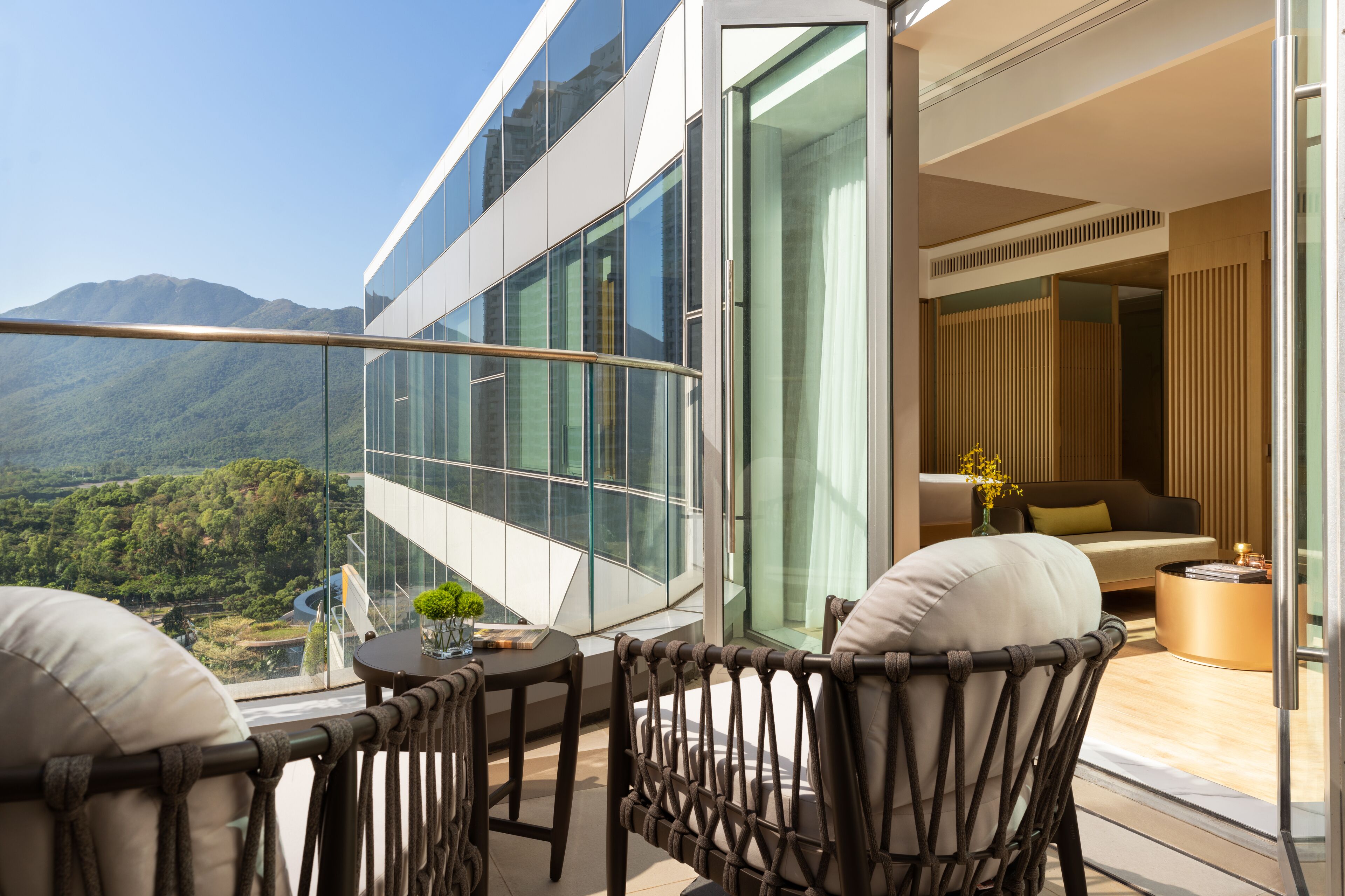 A modern balcony with a glass railing, a wicker chair, a small table with a vase, and a view of a mountainous landscape.