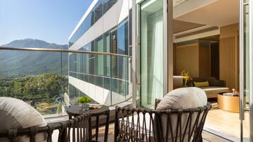 A modern balcony with a glass railing, a wicker chair, a small table with a vase, and a view of a mountainous landscape.