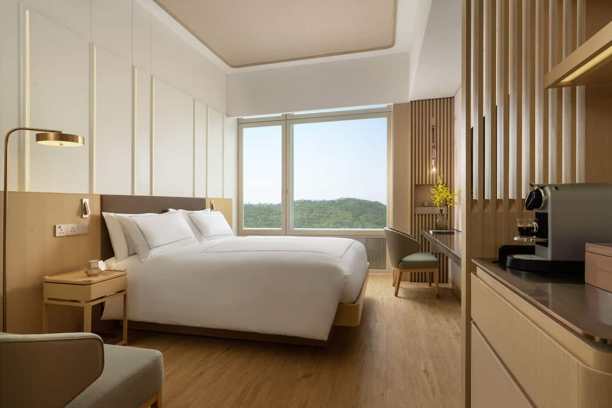 Een moderne hotelkamer met een groot bed, nachtkastje, lamp en uitzicht op groen buiten het raam.