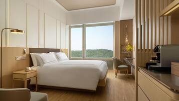 A modern hotel room with a large bed, bedside table, lamp, and a view of greenery through the window.