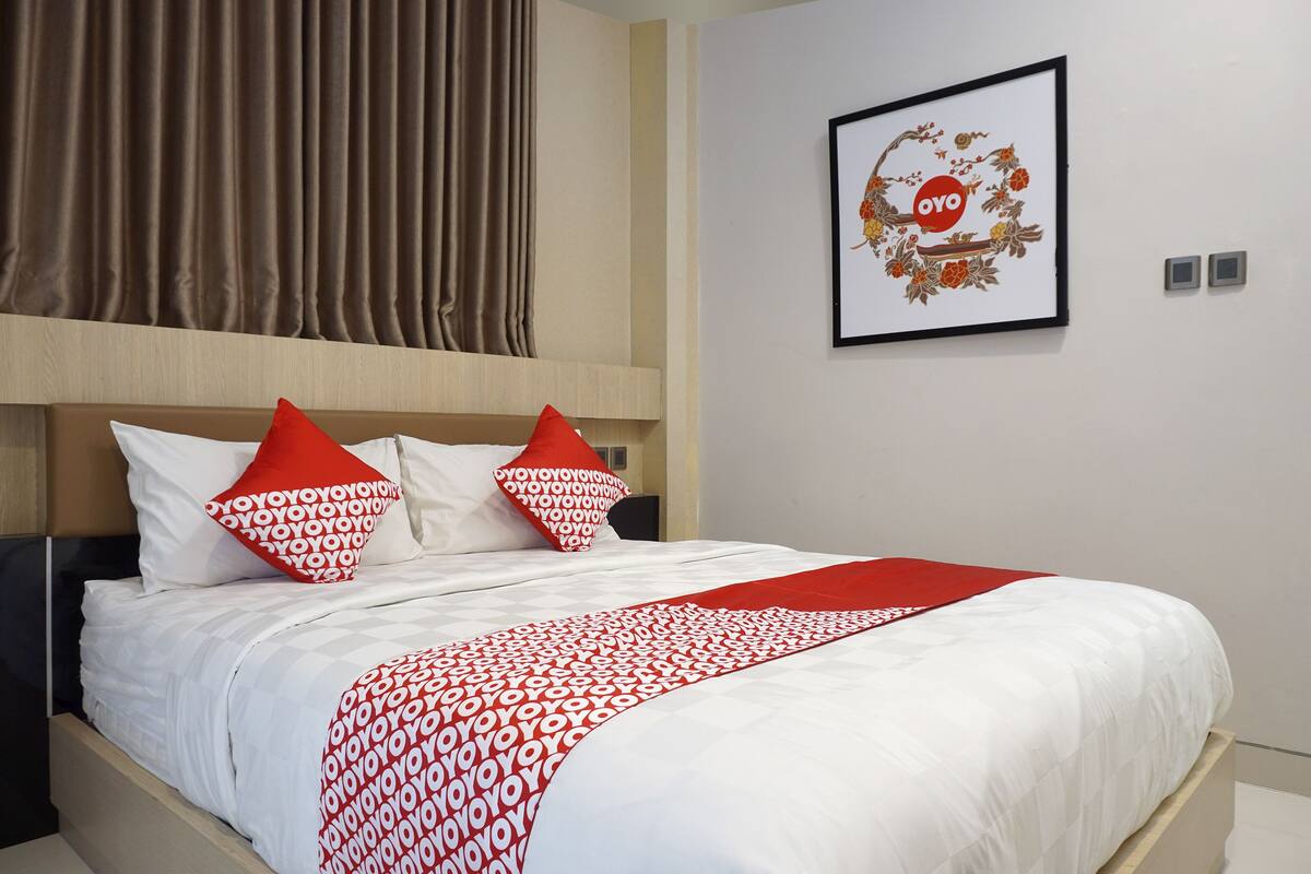 Una cama bien tendida con ropa blanca y detalles rojos, una obra de arte enmarcada en la pared y una cortina entreabierta.