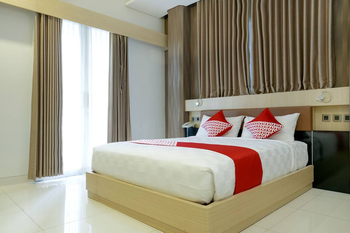 Una habitación de hotel moderna con una cama grande, cabecera de madera y vistas a la ciudad a través de amplios ventanales.