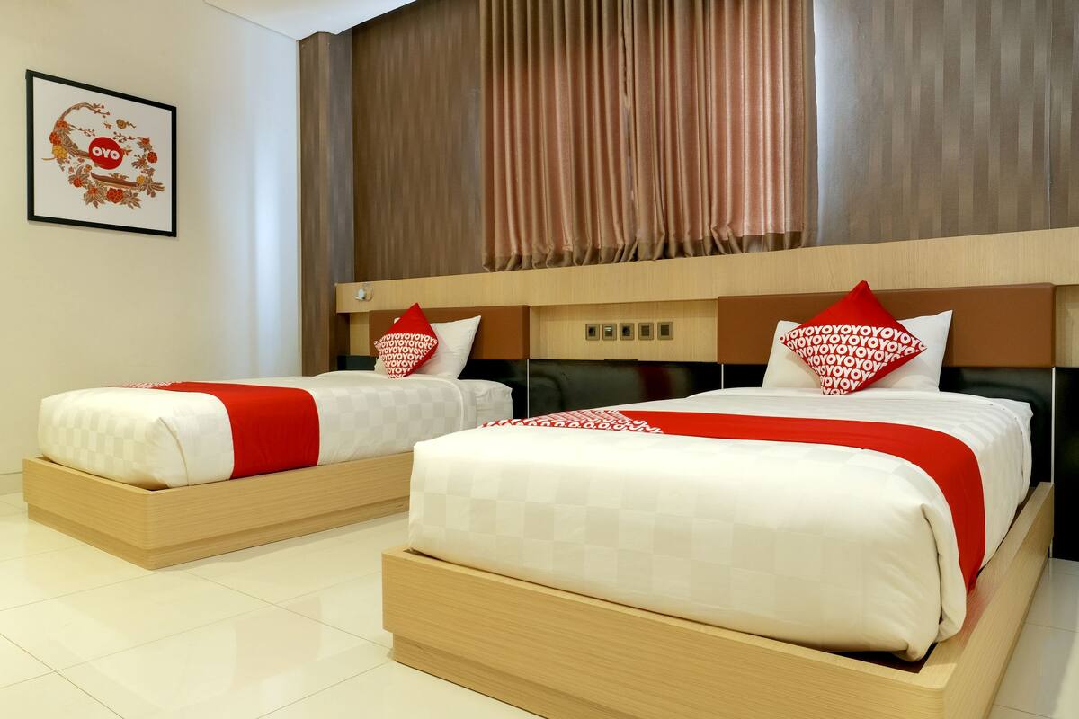 Habitación de hotel con dos camas, cada una con una colcha y almohadas rojas y blancas. La habitación tiene un cabecero de madera y un ventanal con cortinas.