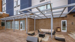 Un espace extérieur aménagé en patio, avec un coin salon couvert, un foyer et un bâtiment en briques en arrière-plan.