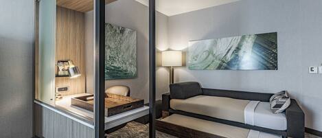 A compact hotel room with a desk, chair, sofa, and artwork on the walls.