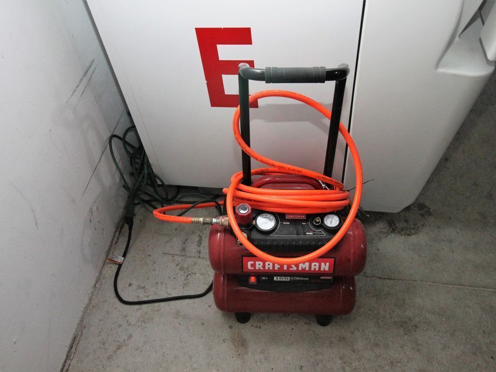 A red Craftsman portable air compressor with an orange hose, placed on a concrete floor next to a white wall with a red 'E' on it.