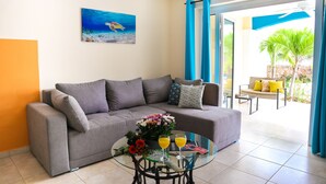 Een woonkamer met een grijze bank, een glazen bijzettafeltje met drankjes en bloemen, een schilderij van een onderwaterlandschap en uitzicht op een terras met een stoel en planten.