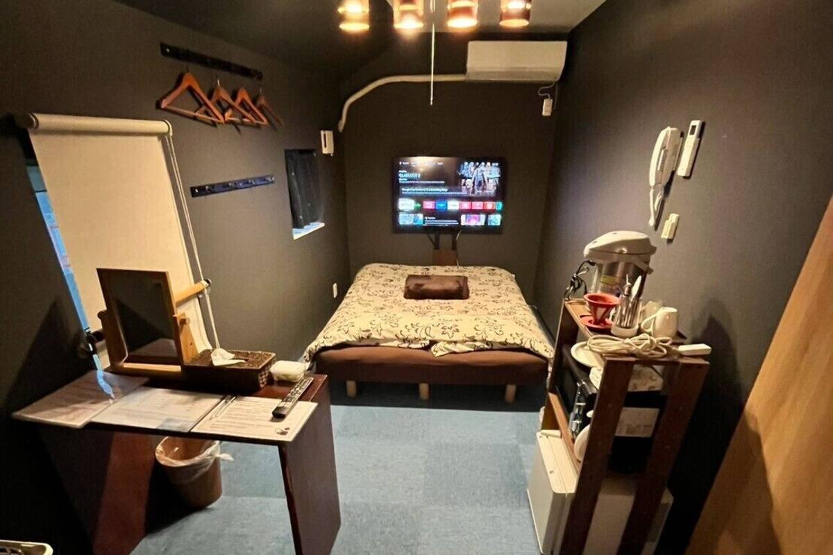 Pieni huone, jossa on sänky, työpöytä, tuoli, pieni pöytä, televisio ja kattovalaisin.