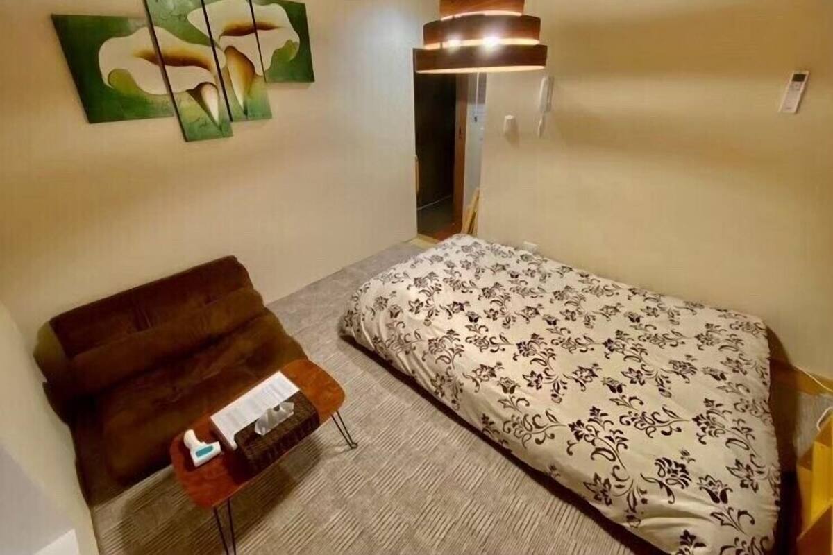 Kompakti hotellihuone, jossa on sänky, sohva, pieni pöytä, jossa on paperipyyhekotelo, sekä seinälle kiinnitetty taideteos.