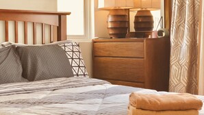 Una cama bien hecha con cubrecama y almohadas estampadas, una mesita de noche de madera con lámpara, y una ventana con cortinas.