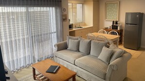 Sala de estar con sofá, una mesa de centro y televisor. Zona de cocina con refrigerador, microondas y lavaplatos.