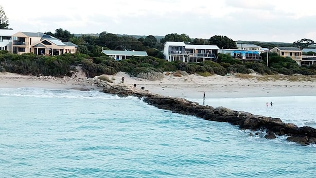 Zona costera con un rompeolas rocoso, playa de arena y varios edificios con balcones que dan al mar.