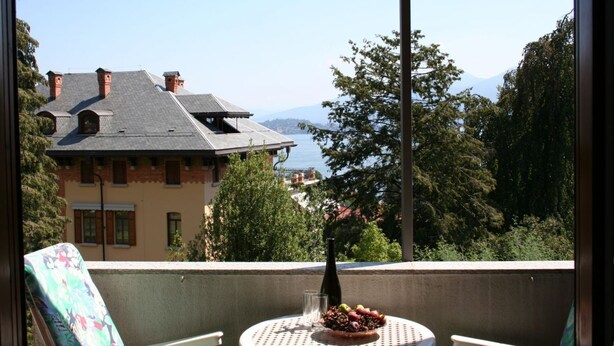 A balcony with a table set for dining, overlooking a building and a mountainous landscape. - Lake view Silvano Apartment (Baveno)