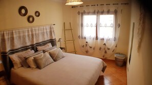 Une chambre à coucher comprenant un lit, des oreillers, une table de chevet, une lampe, une fenêtre avec des rideaux légers et une étagère murale décorative.