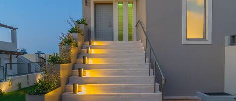 A modern house with a staircase leading to a green door, surrounded by planters and illuminated by warm lighting.