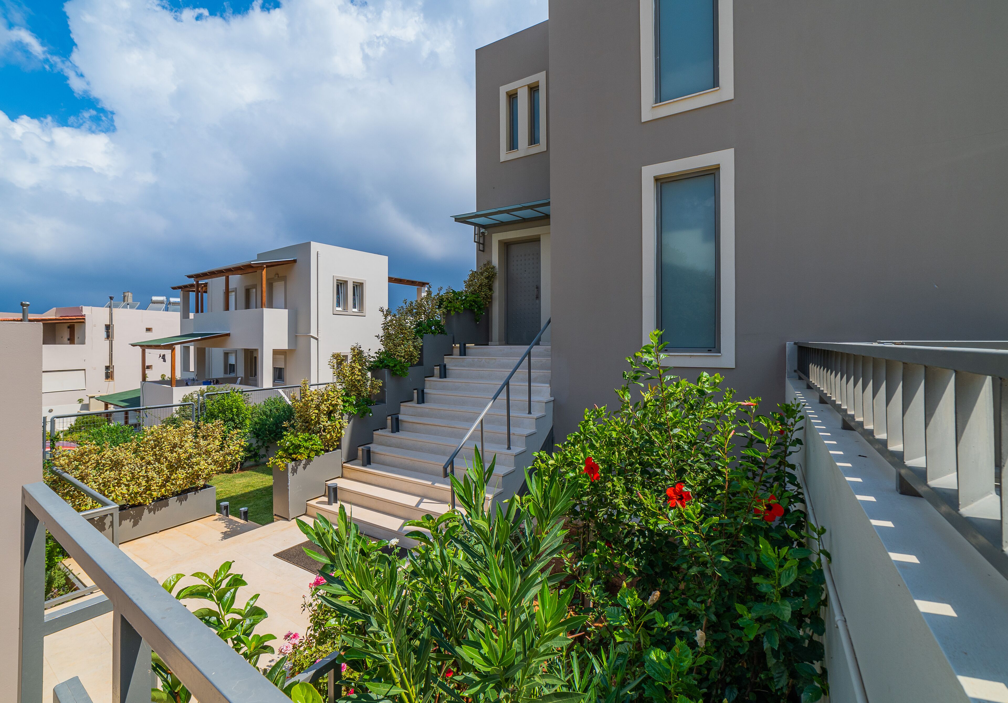 A modern residential building with a staircase leading to the entrance, surrounded by lush greenery and a clear sky.