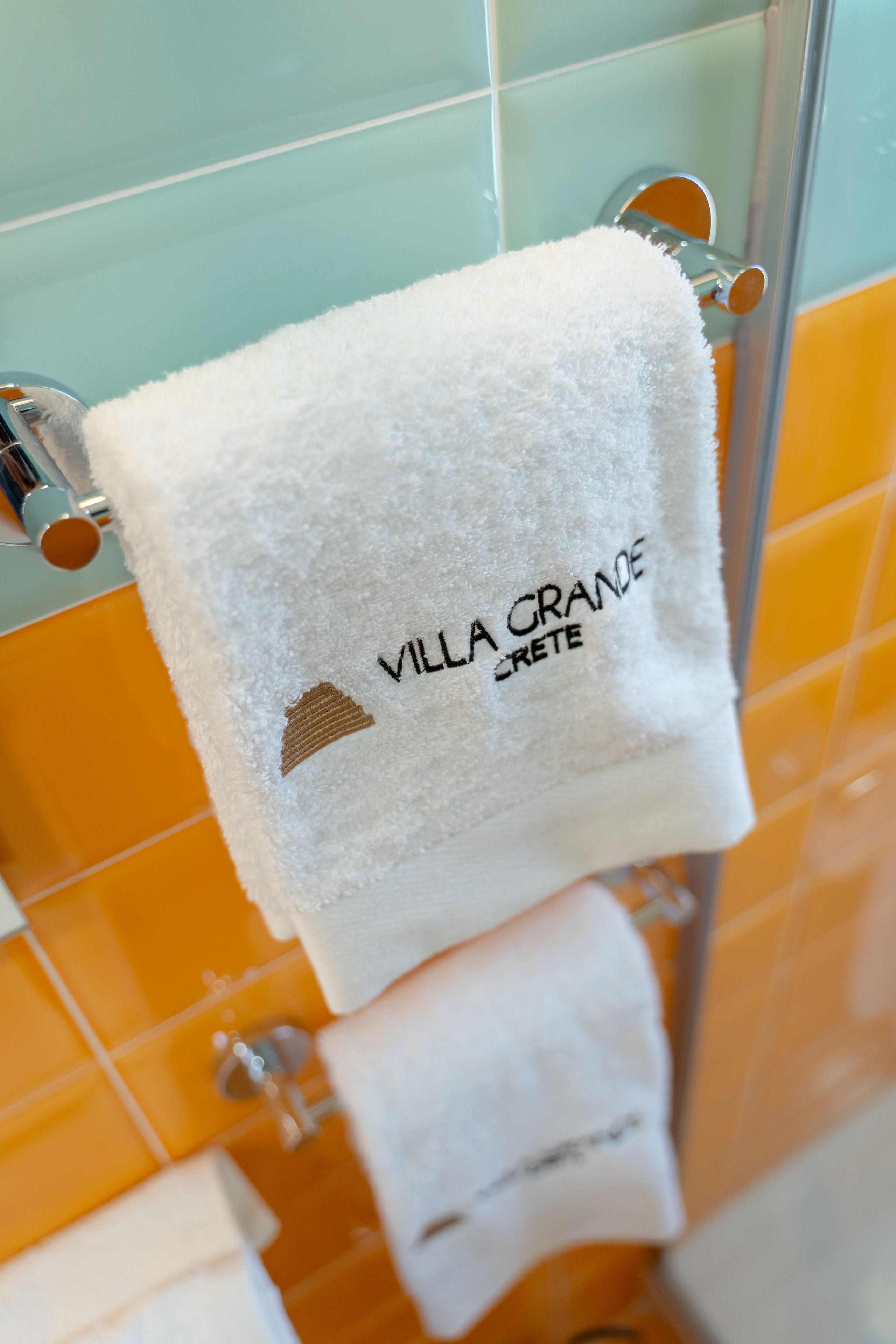A white towel with "VILLA GRANDE CRETE" text and a mountain icon, hanging on a glass shower door in a bathroom with orange tiles.