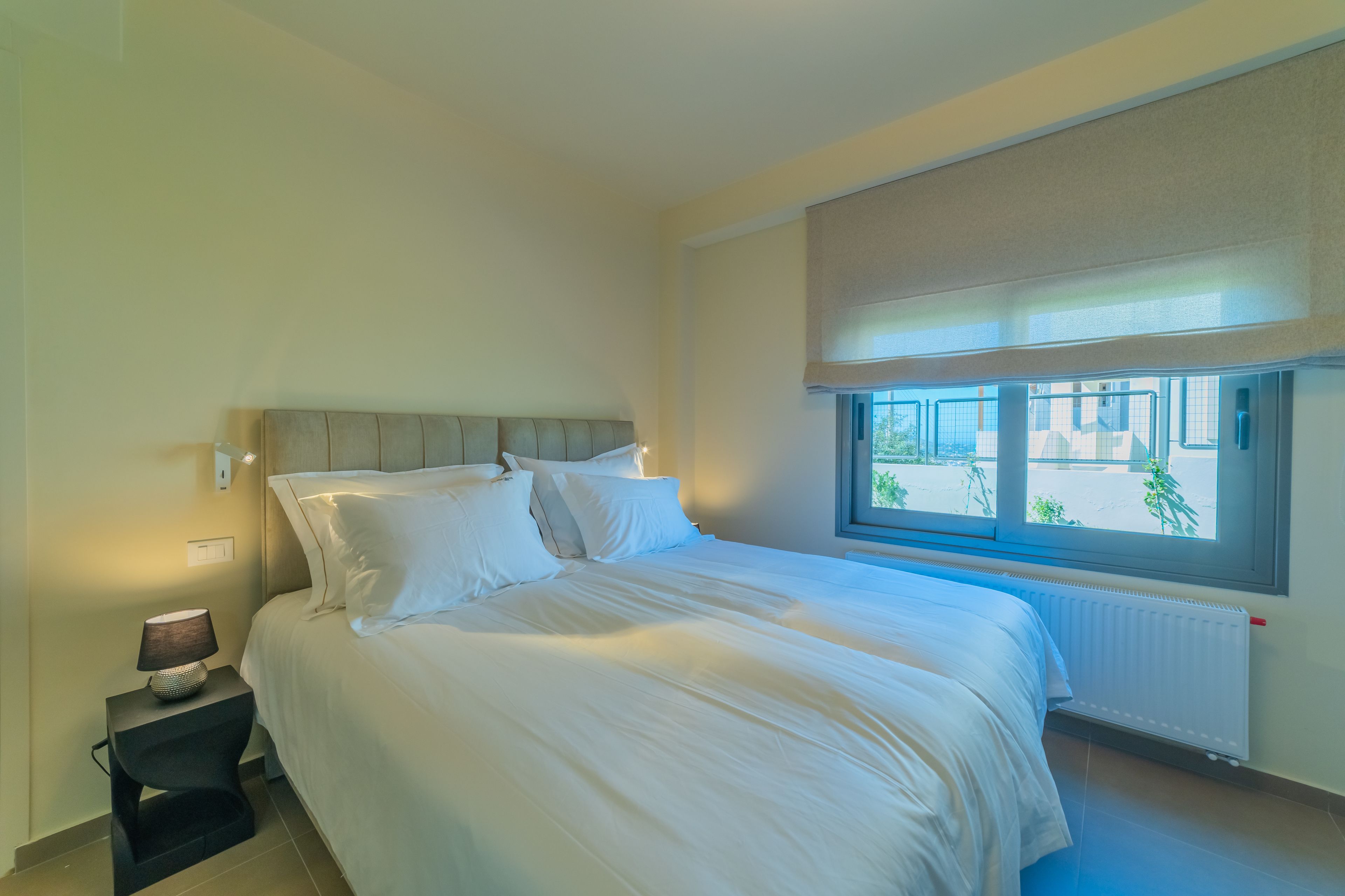 A bedroom with a large bed, a nightstand with a lamp, a window with a view, and a radiator.