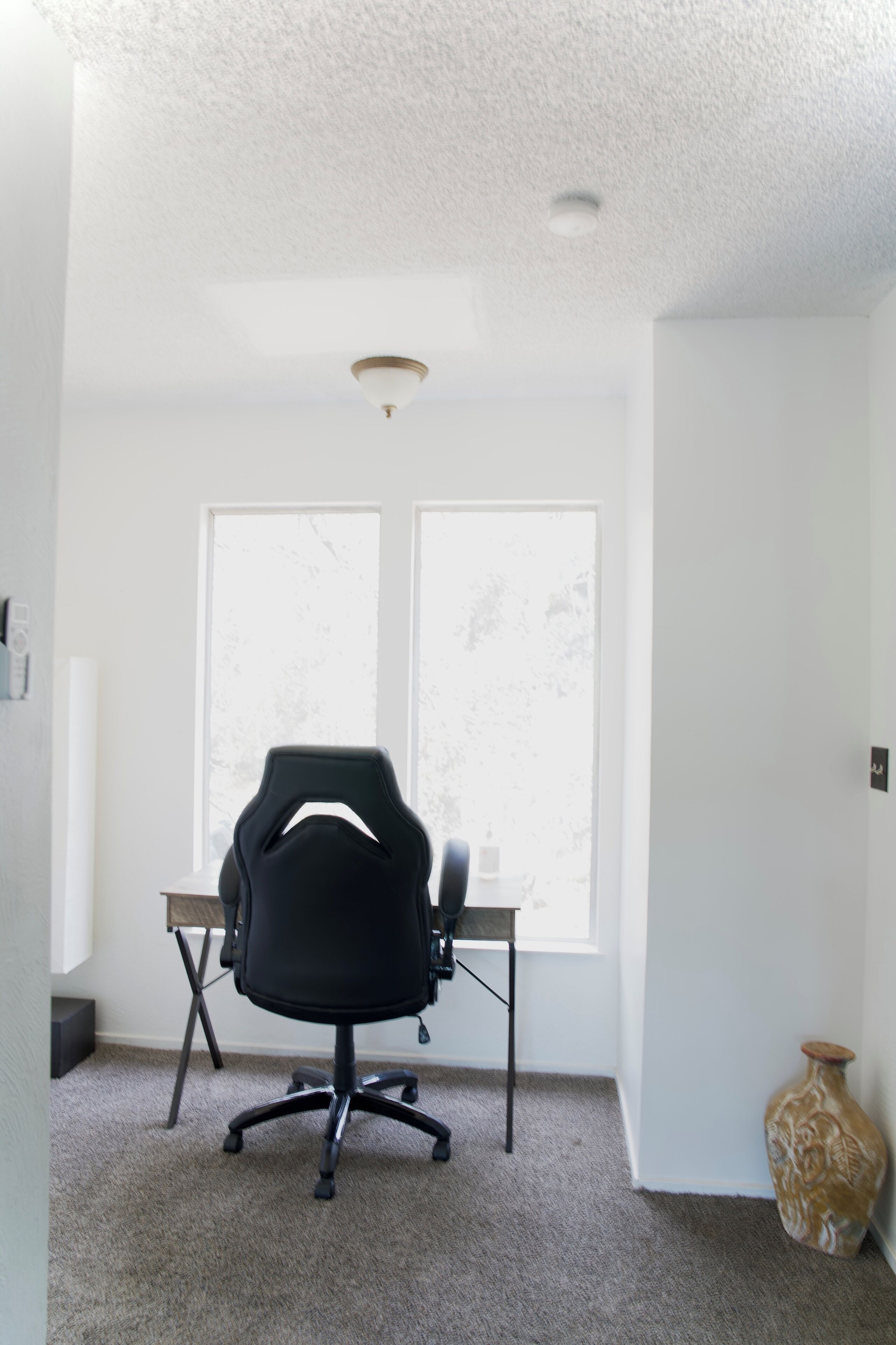 A room with a black office chair, a window, and a vase on the floor.