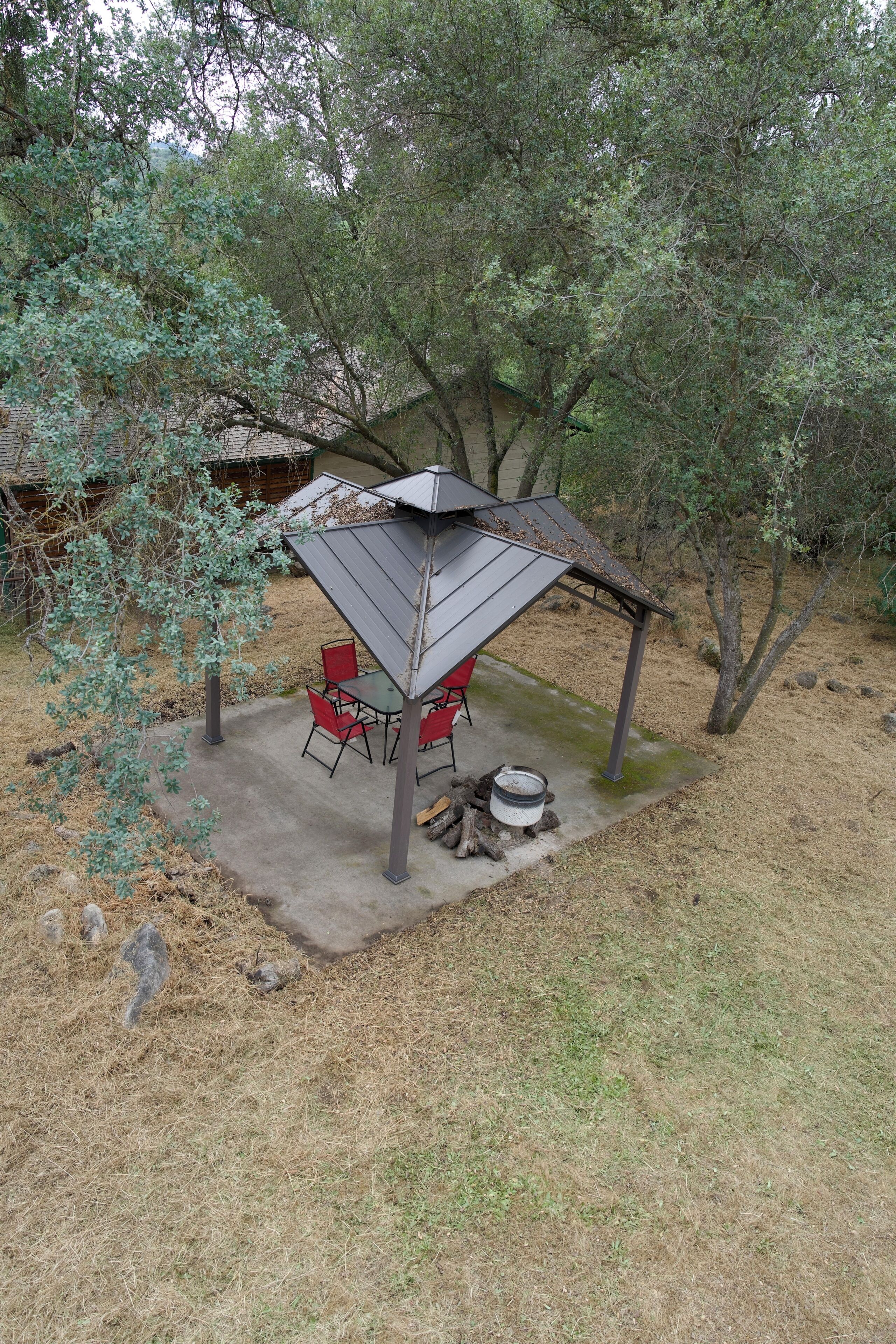A covered outdoor seating area with chairs and a table, surrounded by trees and a fire pit.