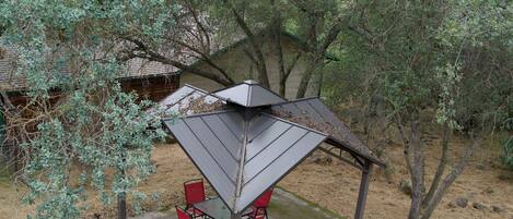 A covered outdoor seating area with chairs and a table, surrounded by trees and a fire pit.