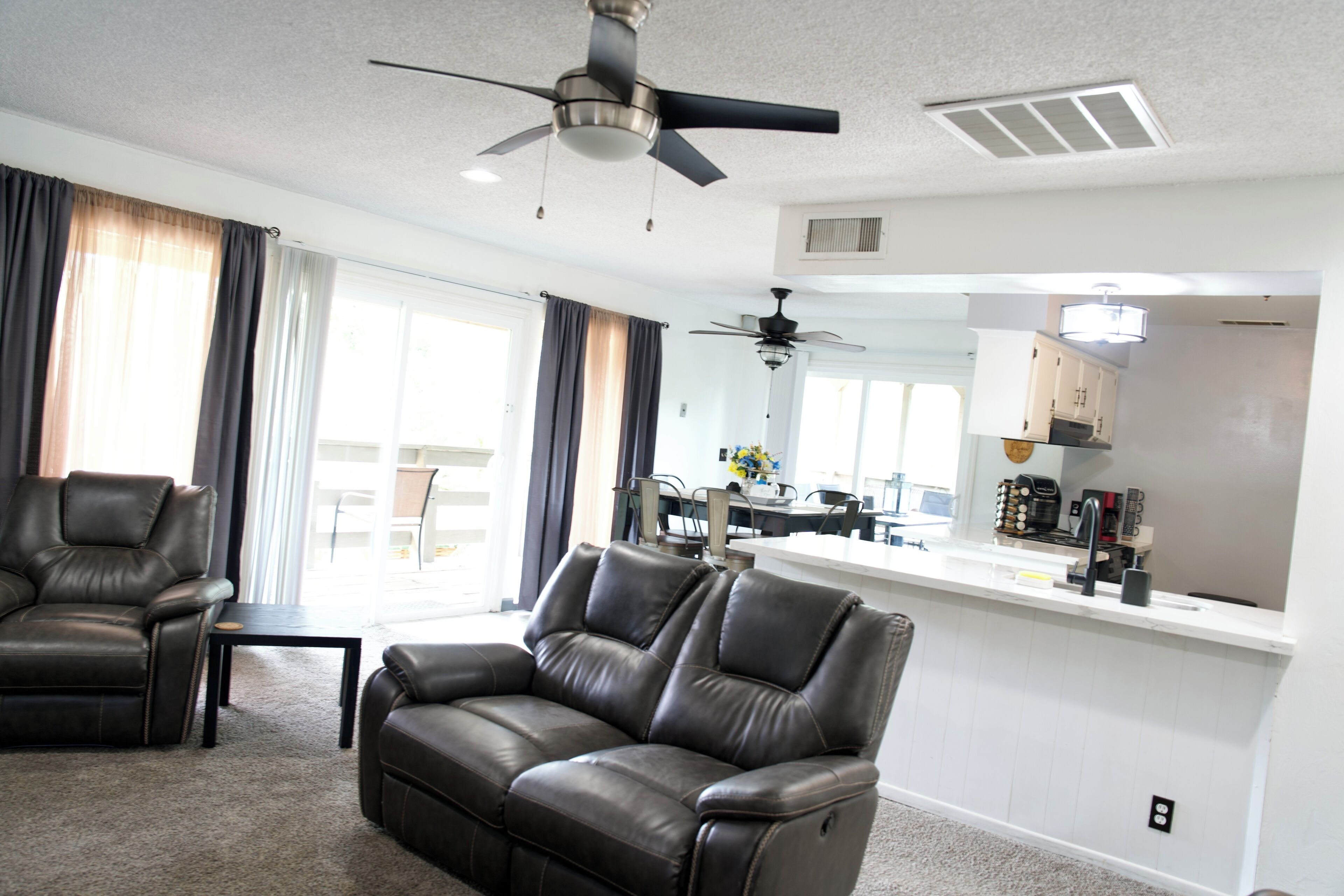 A living room with a sofa, a chair, a ceiling fan, and a kitchen area in the background.