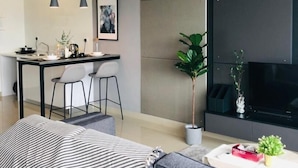 A modern living room with a grey sofa, a coffee table, and a TV. There is a dining area with a table and chairs, and a kitchen area with a stove. - Arte Plus by Poly City (Kuala Lumpur)