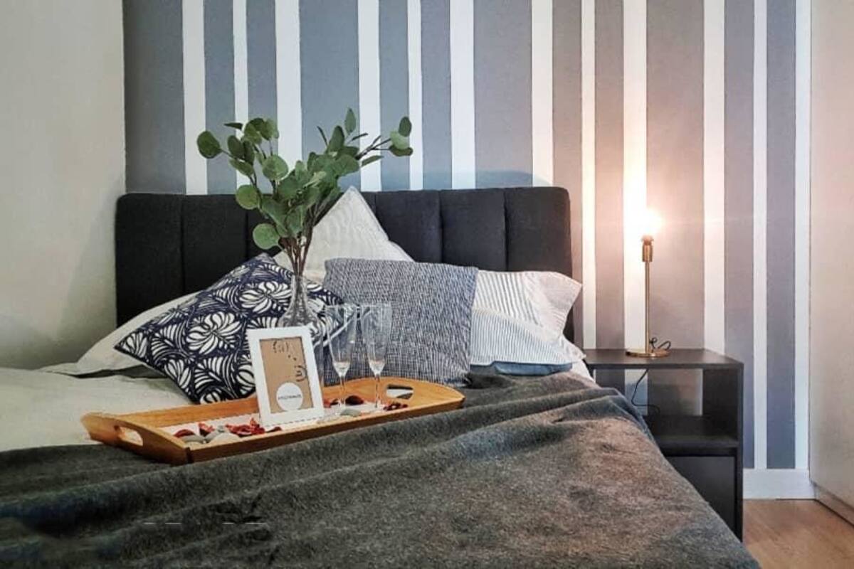 A neatly made bed with a tray of snacks and a wine glass, a bedside plant, and a lamp on a nightstand.