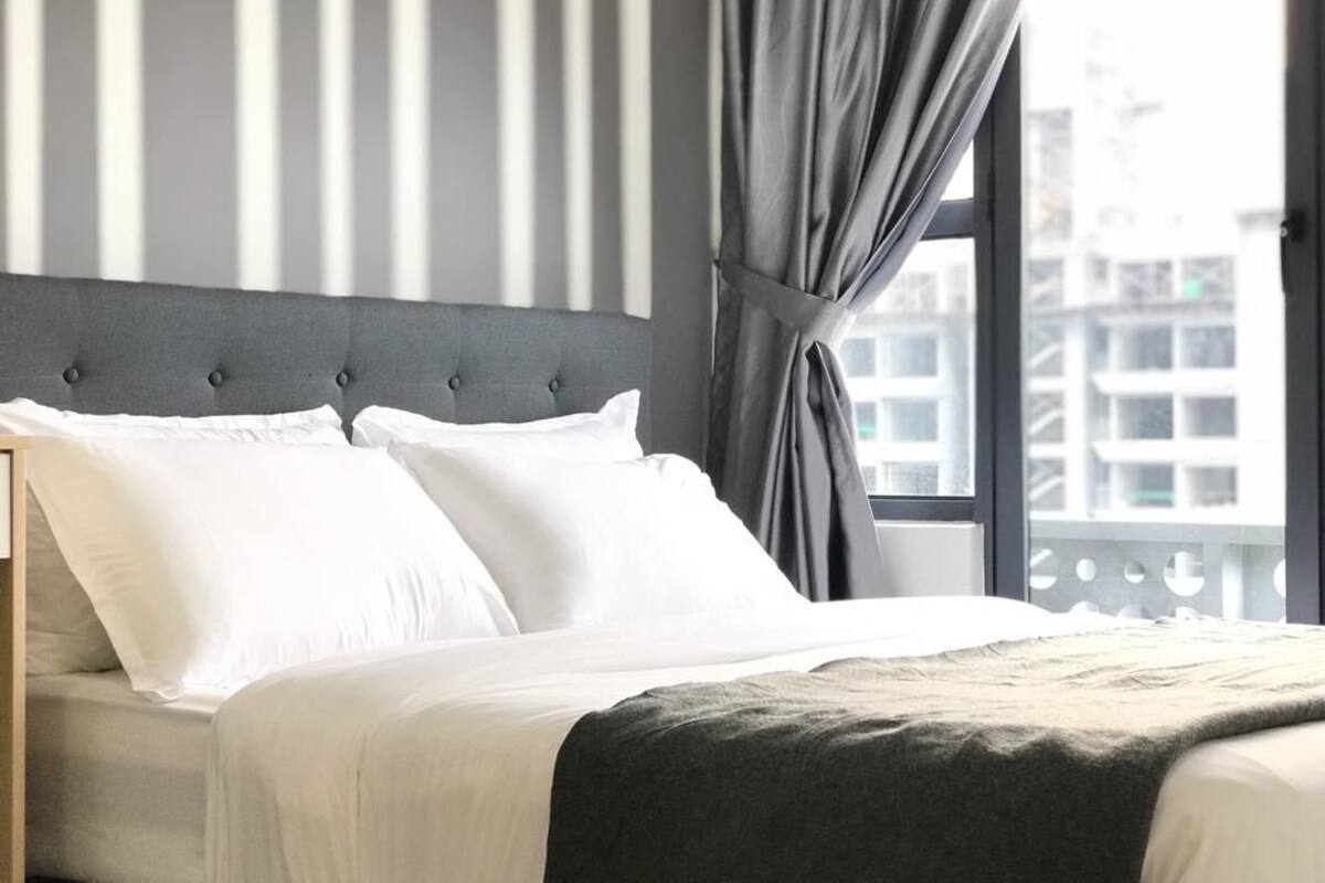 A neatly made bed with white linens and a grey headboard, a window with grey curtains, and a view of buildings outside.