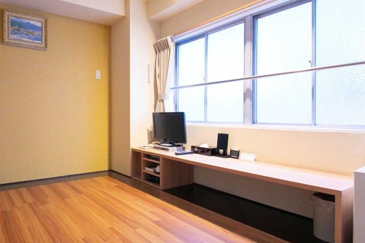 Una habitación con piso de madera, un escritorio con una computadora, un televisor y una ventana con cortinas.