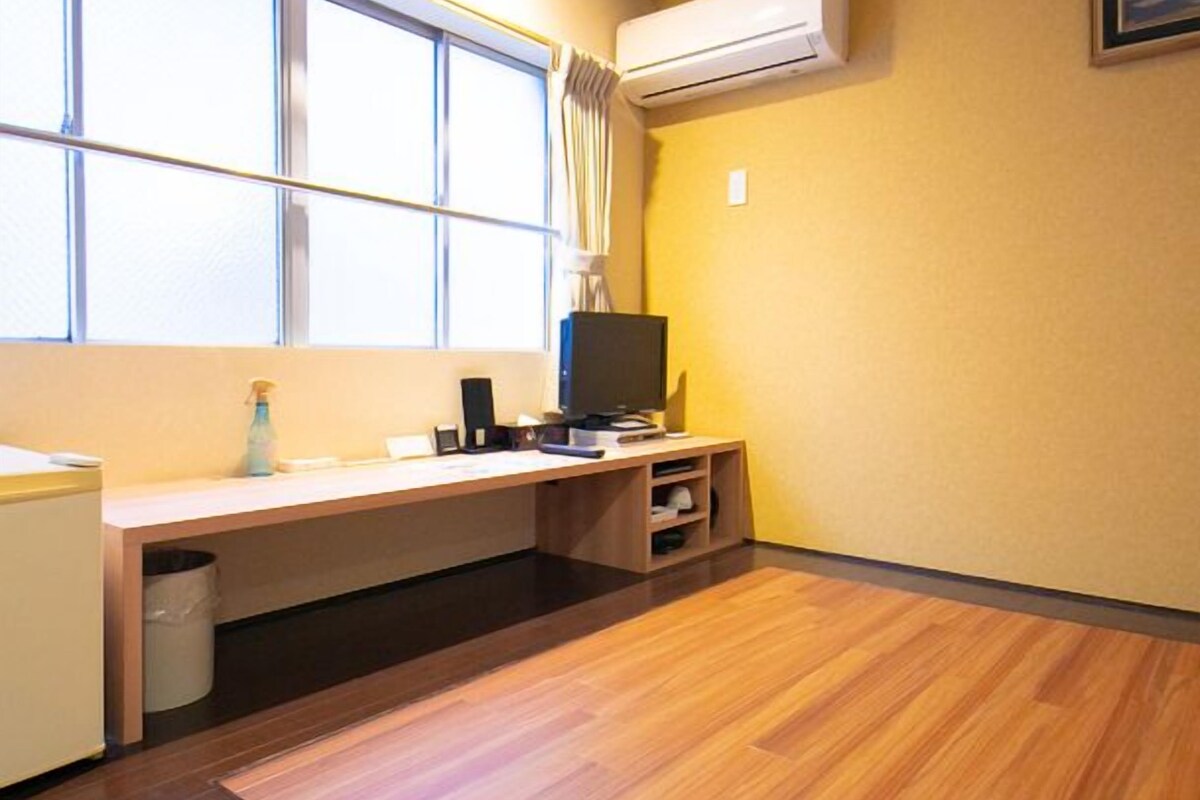 Una habitación con piso de madera, un escritorio con computadora, un televisor y una ventana con cortinas.