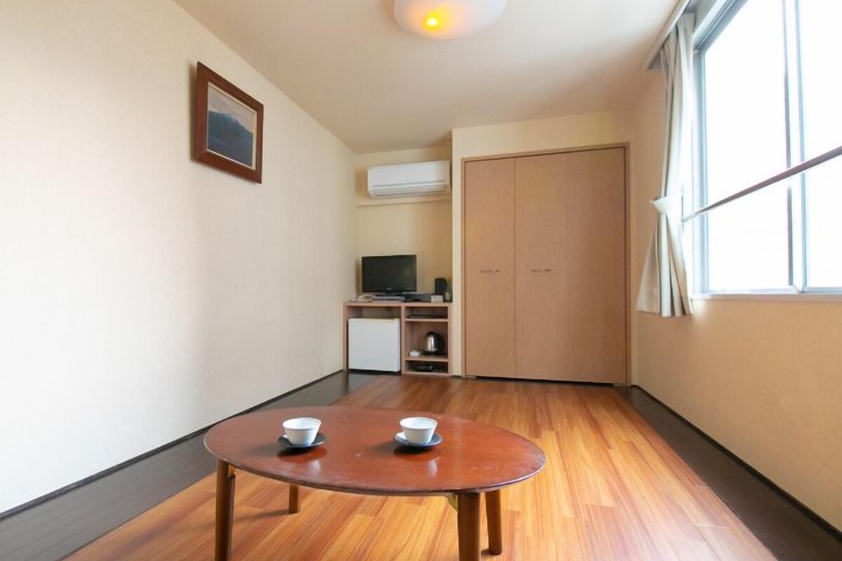 Una habitación con piso de madera, una mesa redonda con dos cuencos, un televisor, una kitchenette y una ventana con cortinas.
