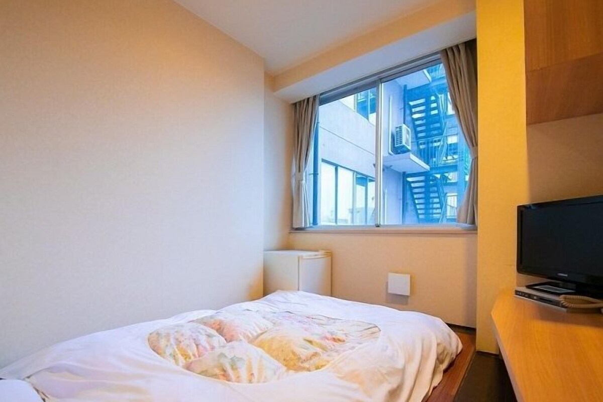 A hotel room with a bed, a TV, a window with a view of buildings, and a radiator.